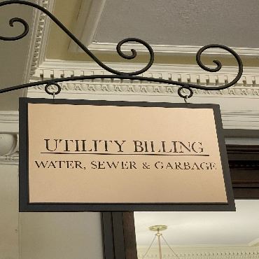 A tan Utility Billing sign hangs from a decorative hook in the main hallway of City Hall.