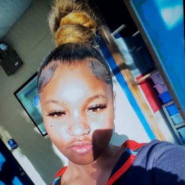 Teen girl taking a selfie. Has brown skin, black hair pulled back, wearing dark hoodie.