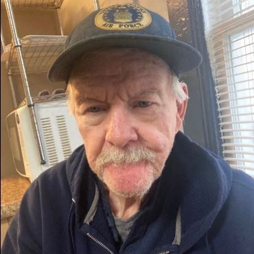 Older white man wearing a baseball-style hat and dark jacket, with white mustache.